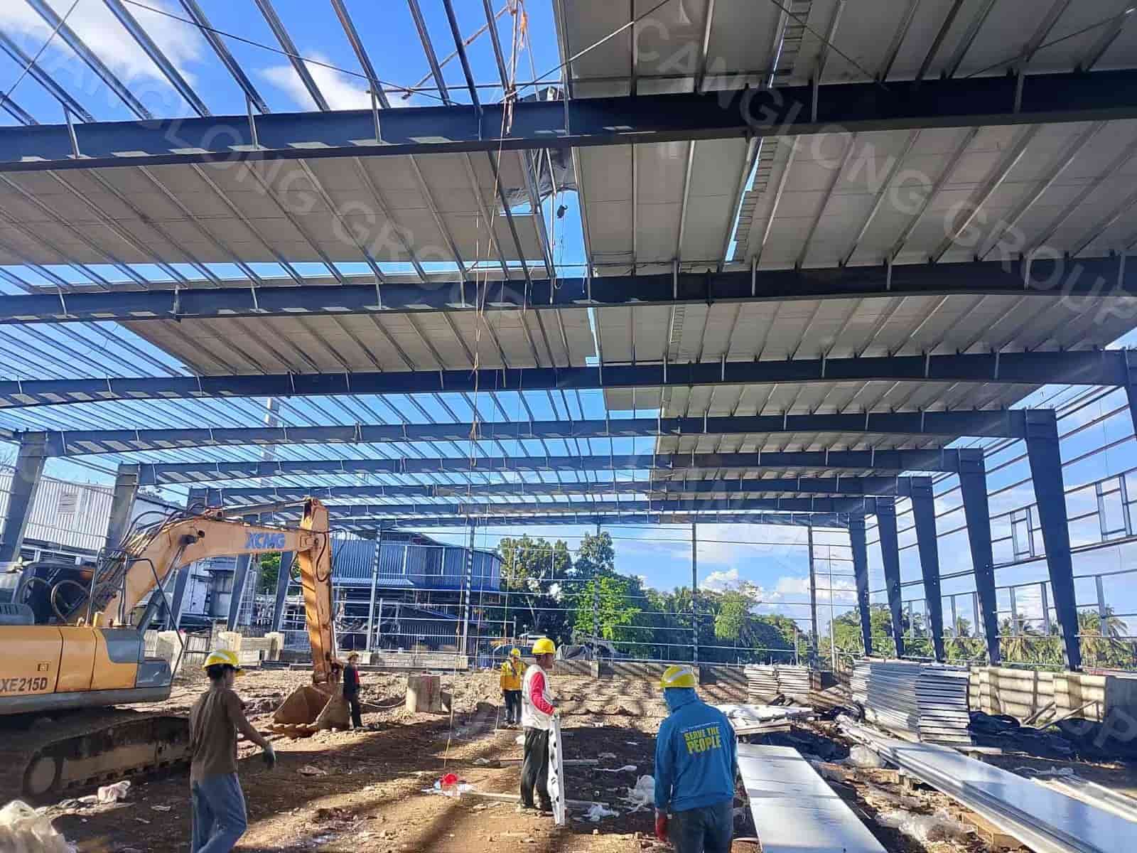 Roof installation underway on a single-slope steel warehouse in Davao, Philippines, with EPS sandwich panels being fixed to the purlins