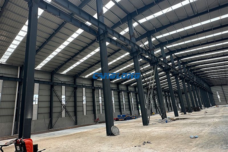 Interior of the Bangkok industrial plant showing the double-span Q355B steel frame, central column line, cross bracing and translucent skylight strips between roof sheets